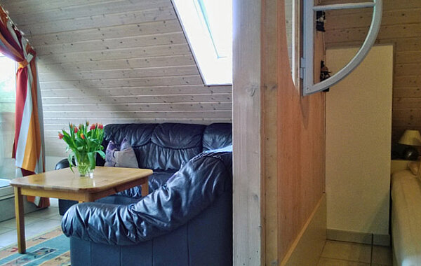 Gemütliches Wohnzimmer mit schwarzer Ledercouch, Holztisch mit Tulpenstrauß und hellem Holzwand-Interieur unter einem Dachfenster. Rechts angrenzender Flur mit einem Bett sichtbar.