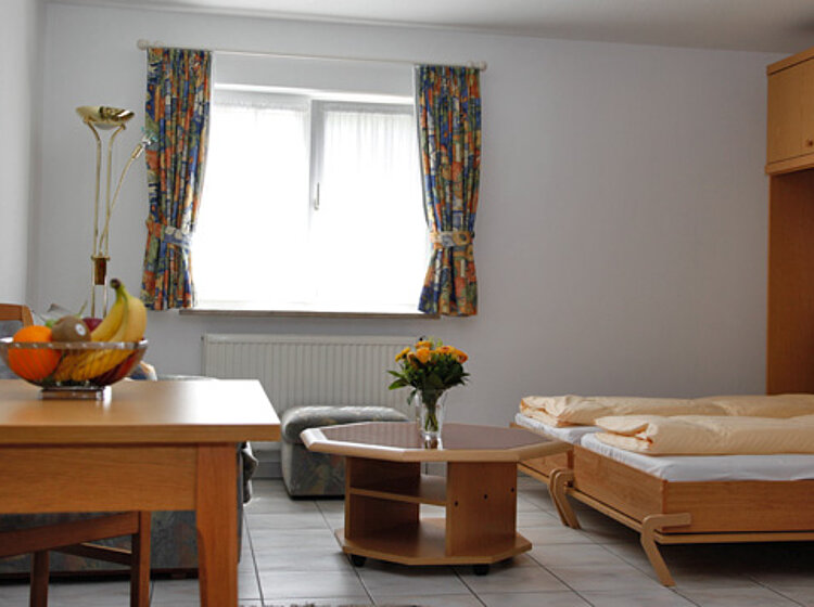 Helles, gemütliches Zimmer mit Einzelbett Helles, gemütliches Zimmer mit Einzelbett, Holzmöbeln, Tisch mit Obstschale und einem kleinen Couchtisch, zwei Fenstern mit bunten Vorhängen und einer Vase mit Blumen.