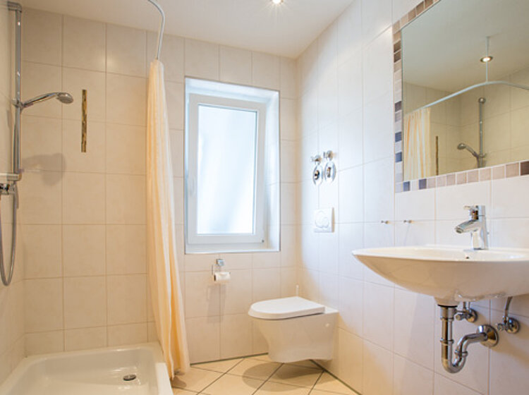 Helles Badezimmer Helles Badezimmer mit ebenerdiger Dusche, Duschvorhang, wandhängendem WC, weißem Waschbecken und großem Spiegel. Ein Fenster sorgt für Tageslicht.