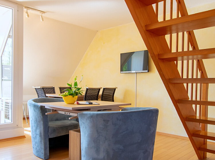 Wohnraum Moderner, heller Wohnraum mit einem Esstisch, blauen Stühlen, einer Pflanze darauf, einer Holztreppe und einem Wandfernseher. Große Glasfenster lassen viel Tageslicht herein.