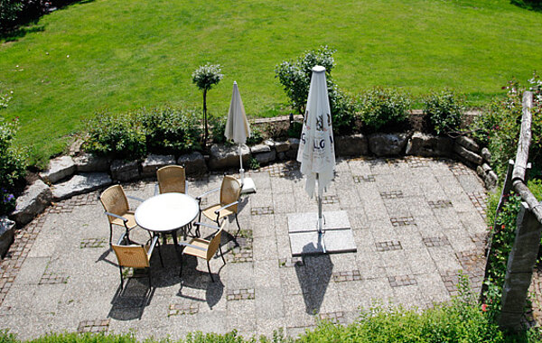 Blick von oben auf eine gepflasterte Terrasse mit einem runden Tisch, fünf Stühlen und zwei geschlossenen Sonnenschirmen, umgeben von Rasen und grünen Pflanzen.