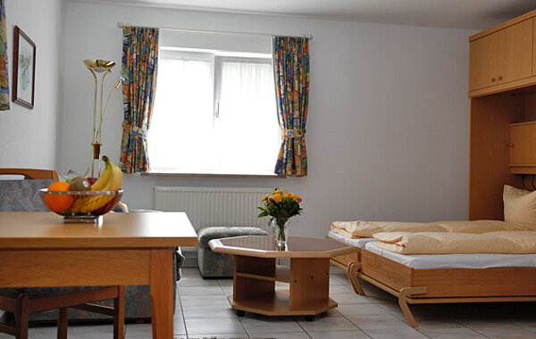 Helles, gemütliches Schlafzimmer mit Einzelbett, Holzmöbeln, einem kleinen Tisch mit Blumen und einer Obstschale auf einem weiteren Tisch vor einem Fenster mit bunten Vorhängen.