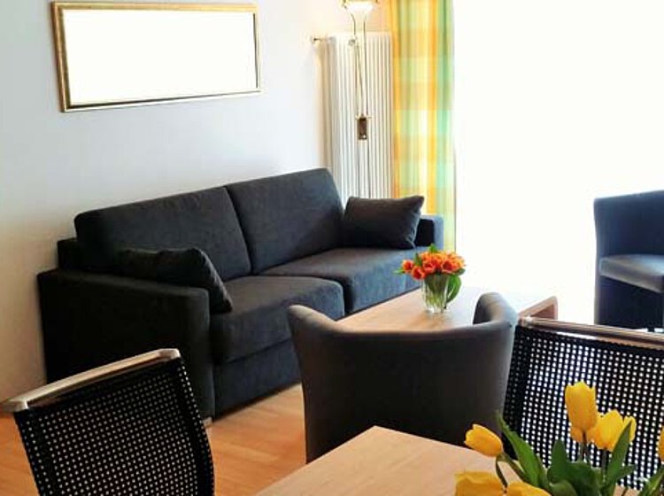 Wohnzimmer mit schwarzem Sofa, zwei Sesseln, einem Tisch mit Blumenvase und hellem Tageslicht durch ein Fenster mit Vorhängen.