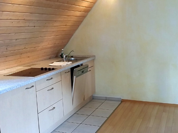 Küchenzeile Kleine Küchenzeile unter einer Dachschräge mit Herd, Spüle, Geschirrspüler und mehreren Schubladen, neben einem offenen Raum mit Holzboden.