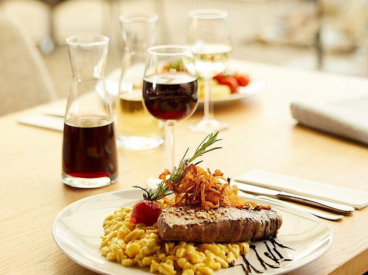 Ein Teller mit einem Stück Rindersteak, Spätzle und gebratenen Zwiebeln, garniert mit einem Rosmarinzweig und Kirschtomate. Im Hintergrund stehen Gläser mit Rot- und Weißwein sowie ein Krug mit Rotwein.