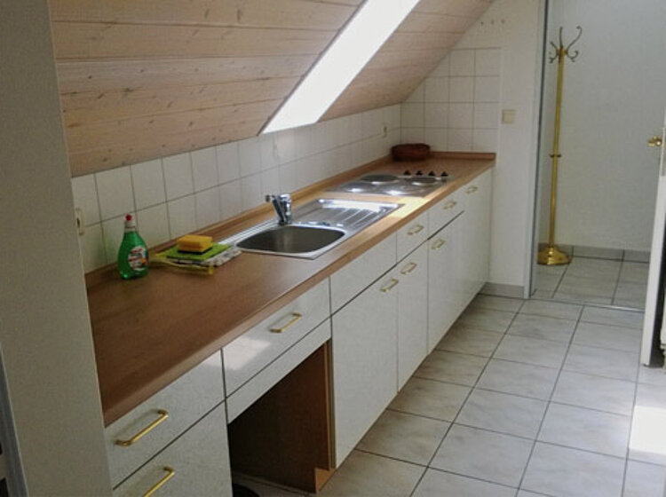 Küche Schmale, schlichte Küche mit Schrägdecke und Dachfenster, langer Holzarbeitsplatte mit Spüle, Herd und mehreren weißen Schränken, heller Fliesenboden, gut beleuchtet durch Tageslicht.