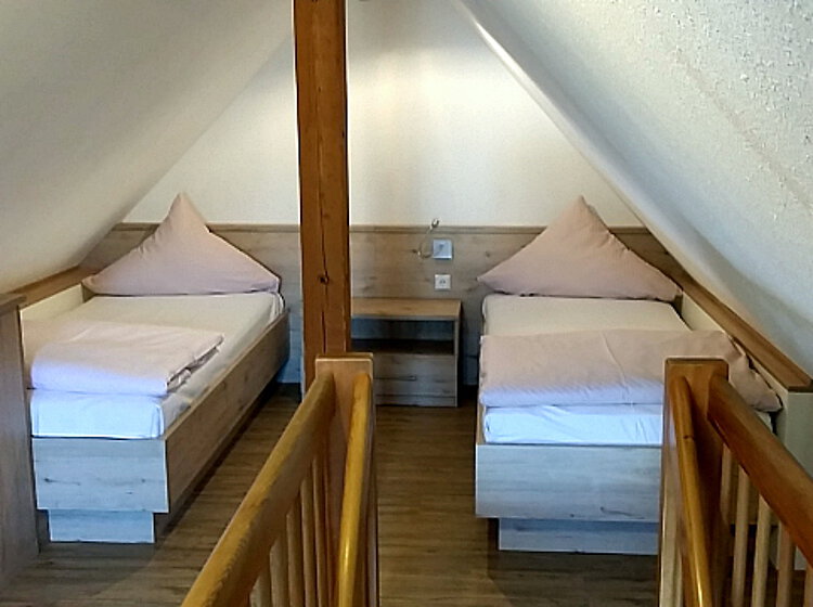 Zwei Einzelbetten Zwei Einzelbetten mit hellen Bettdecken und Kissen stehen unter einer schrägen Holzdecke in einem kleinen Dachzimmer, getrennt durch einen zentralen Holzpfosten und Nachttisch.