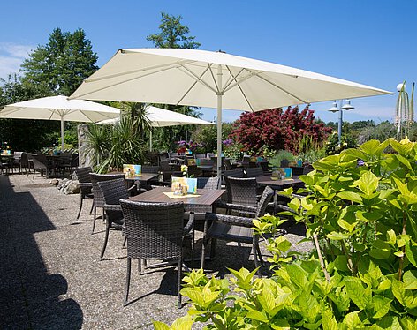 Terrasse Außenbereich eines Cafés mit mehreren Tischen und Stühlen aus Rattan, großen weißen Sonnenschirmen und umgeben von grünen Pflanzen und bunten Blumen bei sonnigem Wetter.
