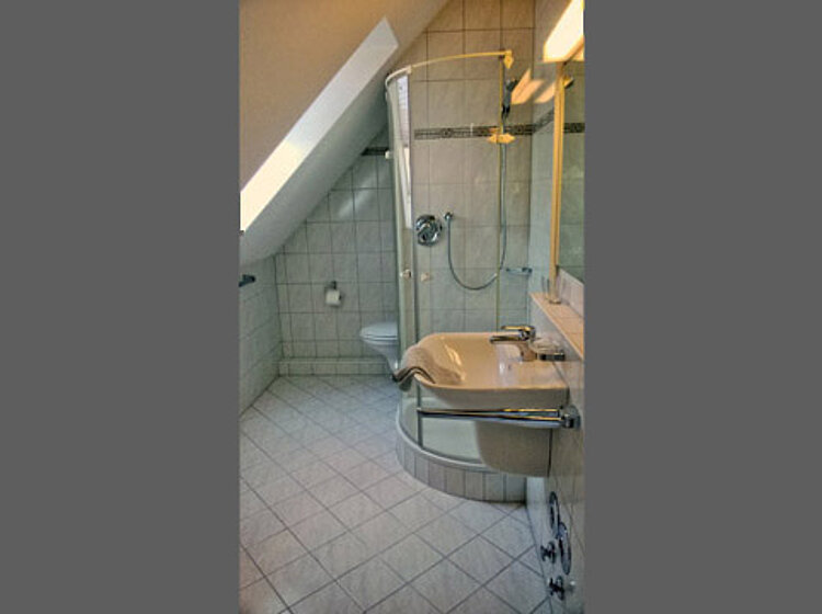 Innenaufnahme eines modernen Badezimmers mit schräger Decke, runder Glasdusche, Waschbecken und Toilette, alle in hellen Fliesen ausgeführt.