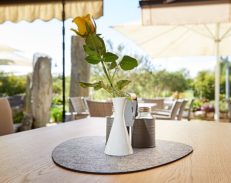 Sonnenterrasse Schlichte weiße Vase mit gelber Rose auf einem Holztisch im Freien, umgeben von Sonnenschirmen und Gartenmöbeln, bei Tageslicht und blauem Himmel.