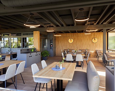 Gastraum Moderner, heller Gastraum mit Holztischen, grauen Stühlen und Sitzbänken. Dezente Beleuchtung, Holzvertäfelung an der Wand und große Fenster sorgen für freundliche Atmosphäre. Tische sind mit kleinen Vasen und Einrichtungsgegenständen dekoriert.