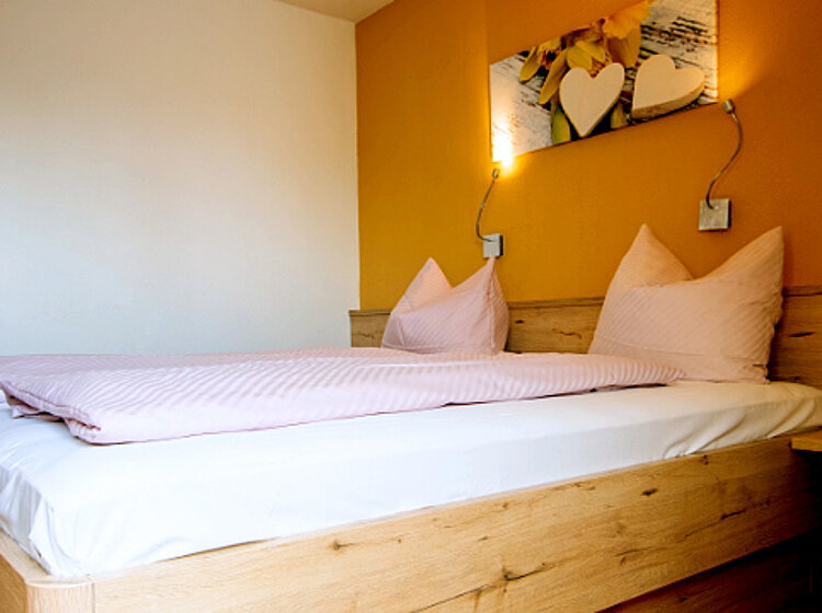 Helles Schlafzimmer Ein helles Schlafzimmer mit einem Doppelbett, rosa gestreifter Bettwäsche und zwei großen Kissen. Die Wand hinter dem Kopfteil ist orange, an der Wand befindet sich ein Bild mit Dekoelementen. Rechts steht ein kleiner Nachttisch.