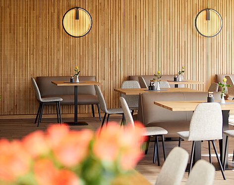 Gastraum Moderner Gastraum mit Holzwandverkleidung, mehreren Tischen und Stühlen, kleinen Vasen mit gelben Blumen auf den Tischen sowie runden Wandlampen, die warmes Licht spenden.