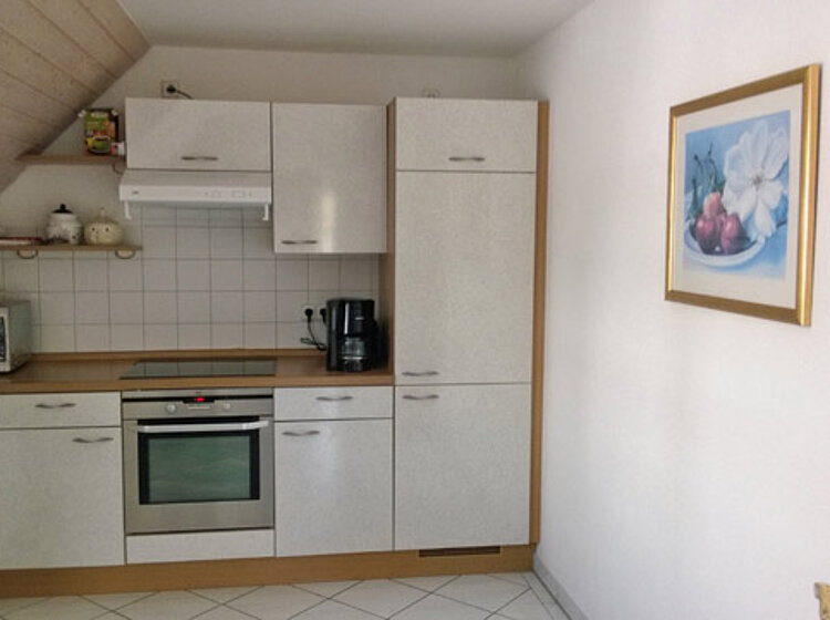 Moderne, helle Einbauküche mit weißen Schränken, Herd, Dunstabzugshaube, Mikrowelle, Kaffeemaschine und Kühlschrank in einer Ecke eines Raumes mit weiß gefliester Wand und Boden.