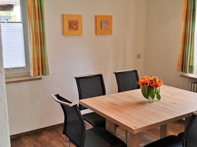 Helles Esszimmer Helles Esszimmer mit einem quadratischen Holztisch, vier schwarzen Stühlen und einem Blumenstrauß in einer Vase in der Mitte des Tisches. Zwei Fenster mit bunten Vorhängen und zwei kleine Bilder an der Wand sind sichtbar.