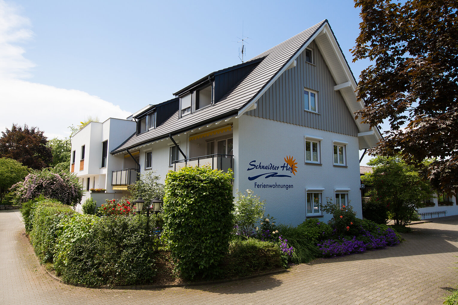Schnaidter Hof Ausenansicht Haus mit Schrägdach und Balkonen, beschriftet mit „Schneider Hof Ferienwohnungen“, umgeben von einem gepflegten Garten mit bunten Blumen und Sträuchern an einem sonnigen Tag.
