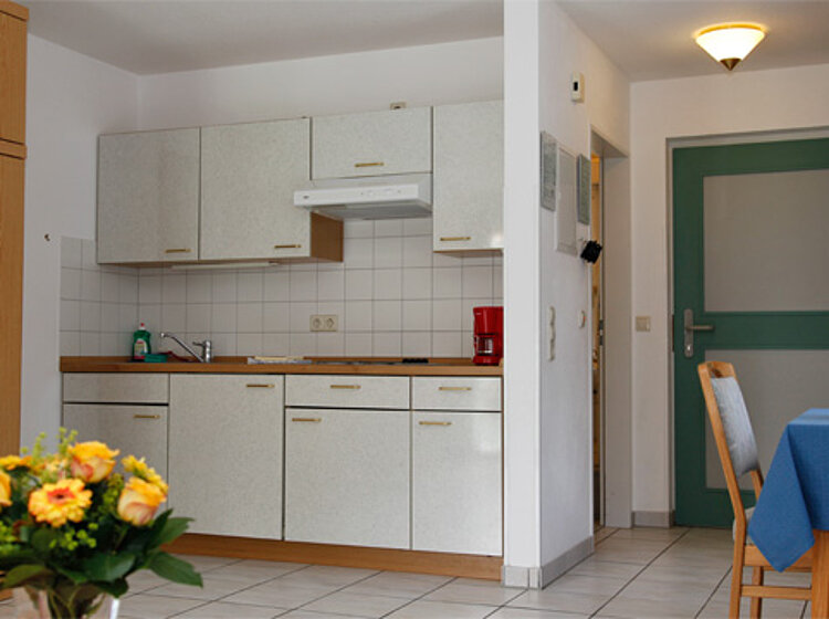 Küchenzeile mit weißen Schränken Kleine Küchenzeile mit weißen Schränken, Spüle, Herd und roter Kaffeemaschine. Im Vordergrund ein Esstisch mit blauem Tischdecken und Blumenschale. Daneben ein Blumenstrauß mit gelben Blumen. Grüner Eingangstür im Hintergrund.