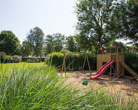 Spielplatz Spielplatz im Garten mit roter Rutsche, zwei Schaukeln und einem Holzfort, umgeben von Büschen, Bäumen und Wiese unter klarem Himmel.