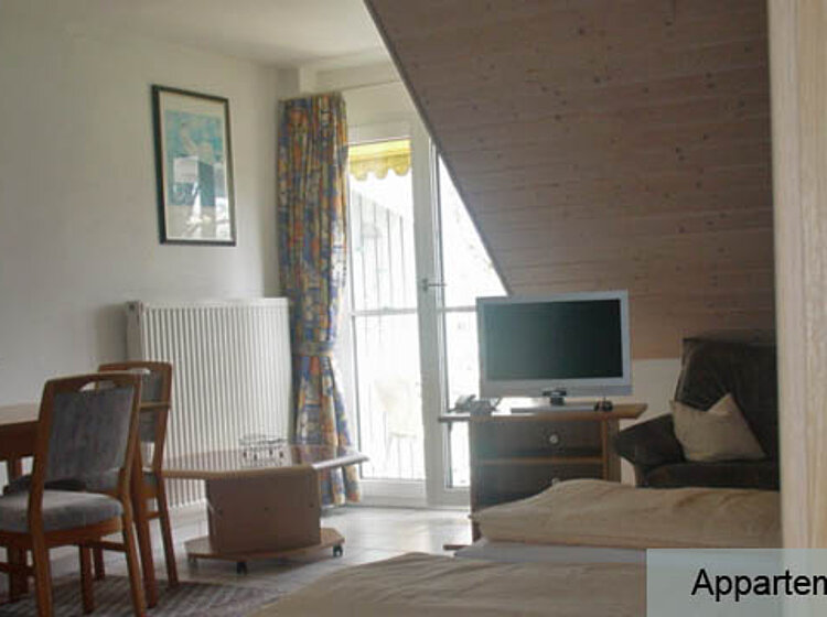 Helles Wohnzimmer Helles Wohnzimmer mit Esstisch und Stühlen, einem Sessel, einem Fernseher auf einem kleinen Tisch und einer Balkontür, die Tageslicht hereinlässt. Die Wände sind weiß, und der Raum wirkt gemütlich und einladend.