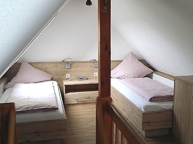 Einzelbetten Zwei Einzelbetten mit hellen Bettdecken in einem kleinen Dachzimmer, getrennt durch einen Holzbalken, mit einem Nachttisch und einem Schrank aus hellem Holz.