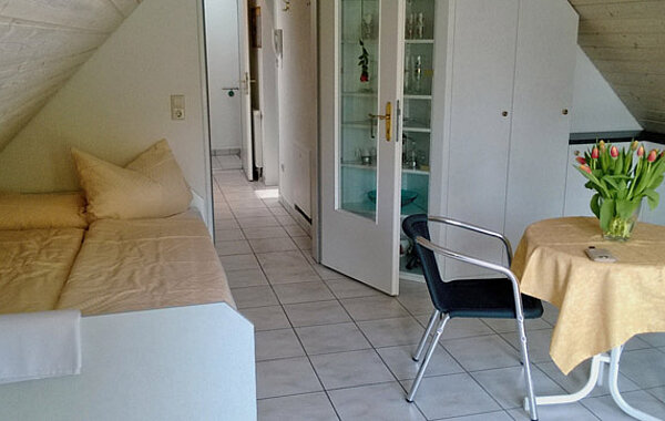 Kleines, gemütliches Zimmer unter Dachschräge mit Einzelbett, Tisch mit gelber Tischdecke, Stuhl und Blumenvase mit roten Tulpen. Heller Fliesenboden und weiße Möbel prägen den Raum.