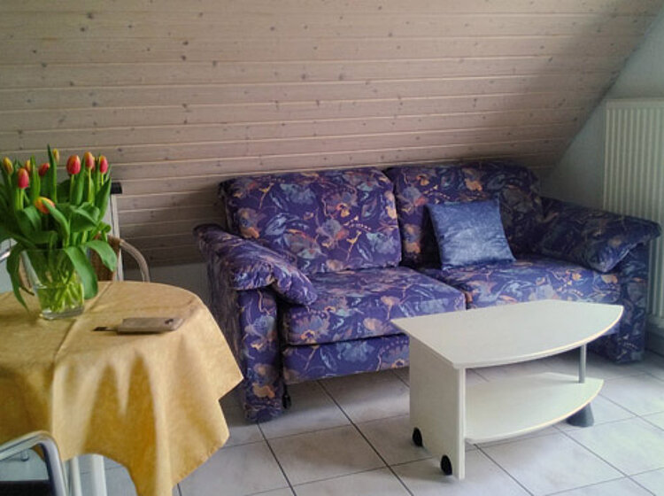 Gemütliches Wohnzimmer Gemütliches Wohnzimmer mit blau gemustertem Sofa, weißem Couchtisch, rundem Tisch mit gelber Tischdecke und Vase mit roten Tulpen.