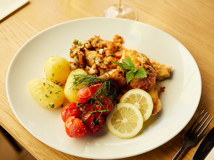 Mittagessen Ein Weißer Teller mit gekochten Kartoffeln, gebratenen Kirschtomaten, gebratenem Fischfilet, garniert mit Zitronenscheiben und frischen Kräutern.
