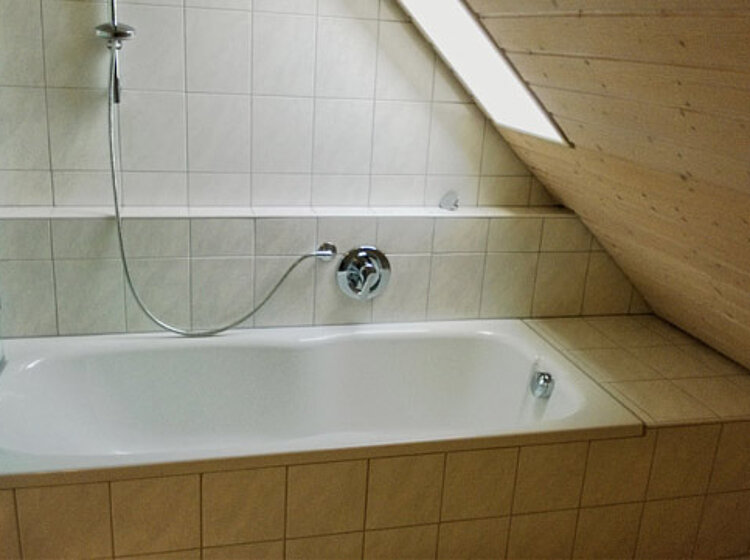 Badewanne Ein weiß eingebautes Badewanne unter einer Dachschräge mit hellen Fliesen an Wänden und Armaturen, inklusive verstellbarer Handbrause.