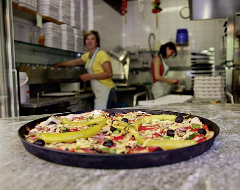 Pizzastation Auf einer Arbeitsfläche in einer Pizzeria liegt eine frisch gebackene Pizza mit Paprika, Oliven und Käse. Im Hintergrund arbeiten zwei Pizzabäckerinnen.