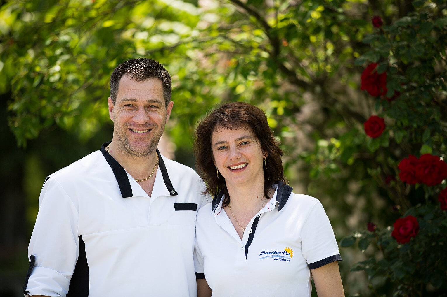 Ein lächelndes Paar in weißer Arbeitskleidung steht unter einem Baum mit roten Rosen, vermittelt Freundlichkeit und Professionalität.