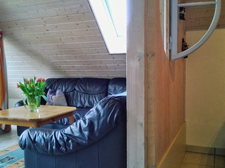 Gemütliches Wohnzimmer mit schwarzer Ledercouch, Holztisch mit Blumenstrauß, Fenster im Dach und angrenzendem Schlafzimmer mit Doppelbett.