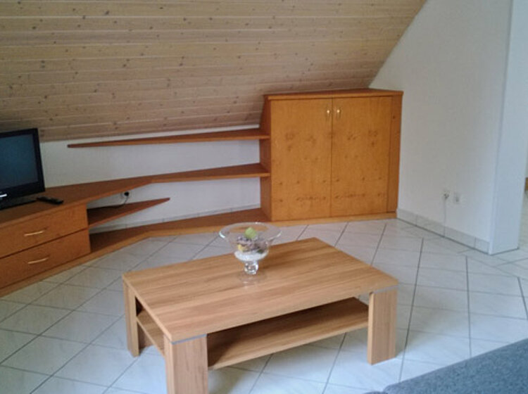 Helles Wohnzimmer mit schräger Holzdecke, Fliesenboden, einem kleinen Fernseher auf einem niedrigen TV-Möbel, einem kleinen Holztisch mit Glasvase darauf und einer kleinen Schrankwand an der Wand. Im Hintergrund ist eine offene Küchenecke sichtbar.