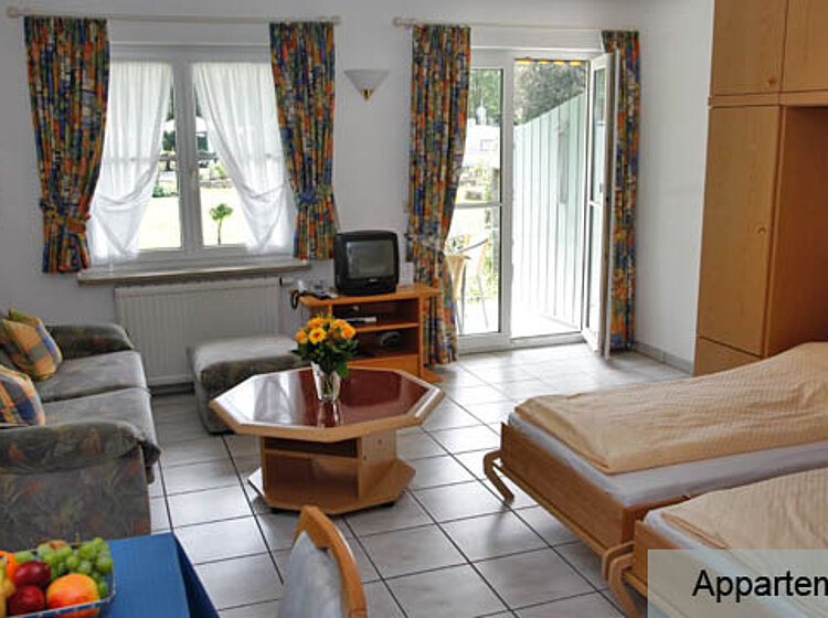 Helles Wohnzimmer Helles Wohnzimmer mit zwei Einzelbetten, Sofa, kleinem Fernseher, Hexagon-Tisch mit Sonnenblumenstrauß und Zugang zu einem Balkon. Fenster mit gemusterten Vorhängen für gemütliche Atmosphäre.