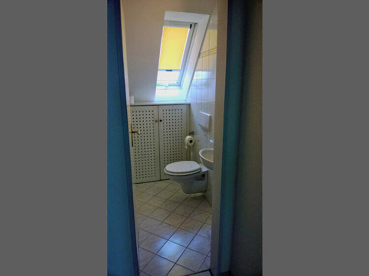 Blick in ein kleines Badezimmer mit Toilette und Waschbecken unter einem Dachfenster. Das Zimmer ist schlicht und hell gefliest.