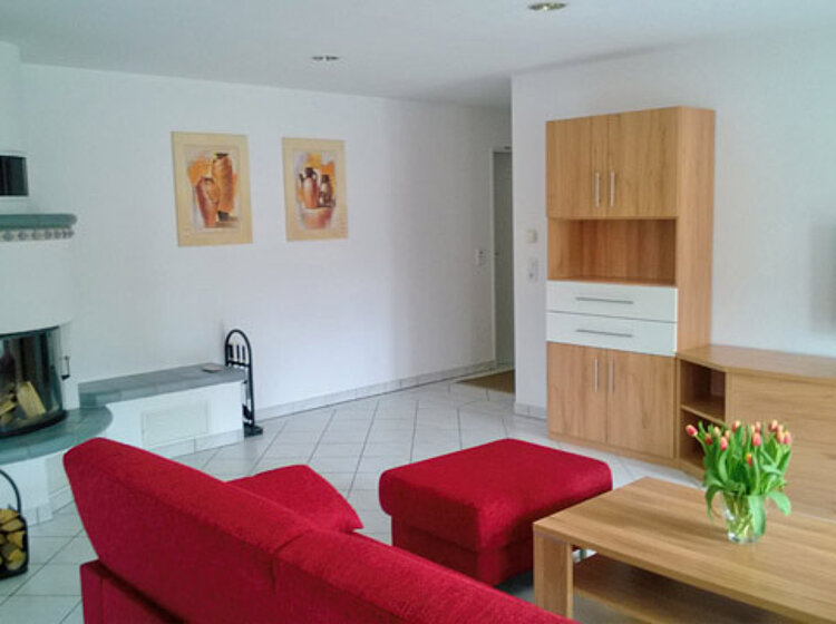 Helles Wohnzimmer mit rotem Sofa und Hocker, einem Holztisch mit Tulpen, Kamin mit Holzlager, weißen Fliesen und Wandregalen sowie Fernseher an der Wand.