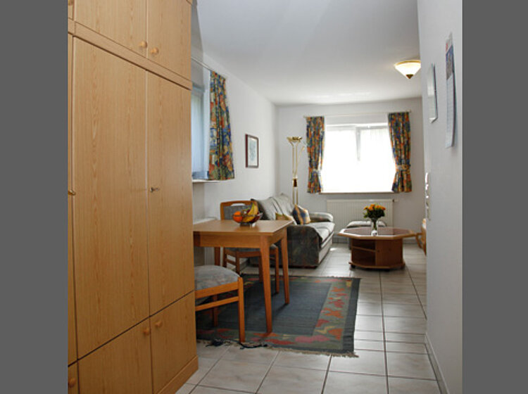 Wohnzimmer mit Esstisch Blick in ein schlicht eingerichtetes Wohnzimmer mit Esstisch, Stühlen, Sofa und Couchtisch vor einem Fenster mit bunten Vorhängen. Teppich und Stehlampe ergänzen die gemütliche Atmosphäre.