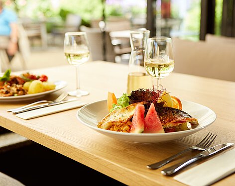 Restauranttisch mit Frühstück Auf einem Restauranttisch stehen zwei Teller mit abwechslungsreichen Mahlzeiten, jeweils begleitet von einem Glas Weißwein und Besteck, die eine gemütliche Essenssituation in einem hellen, einladenden Ambiente zeigen.