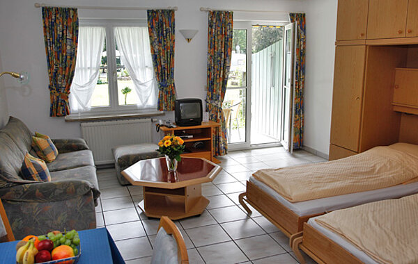 Helles Wohnzimmer mit Sofa, Couchtisch, Fernseher, Esstisch mit Obstschale und zwei Einzelbetten, hellen Vorhängen und Zugang zu einem Balkon oder Garten.