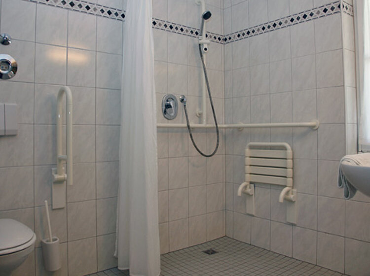 Barrierefreies Badezimmer Barrierefreies Badezimmer mit ebenerdiger Dusche, Duschvorhang, Haltegriffen an der Wand und neben der Toilette sowie einem Waschbecken, alles in hellen Fliesen gehalten.
