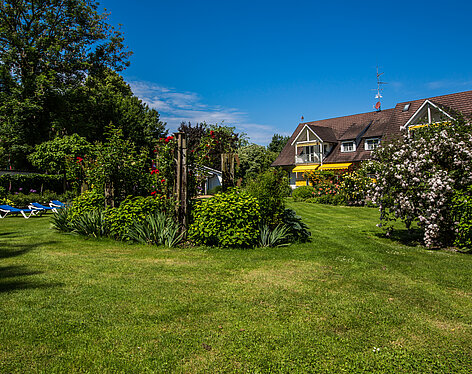 Garten Großzügiger, gepflegter Garten mit grünen Rasenflächen, blühenden Sträuchern und Rosen. Im Hintergrund ein Haus mit braunem Dach und gelben Markisen. Blaue Liegestühle stehen links auf dem Rasen, blauer Himmel mit einigen Wolken darüber.