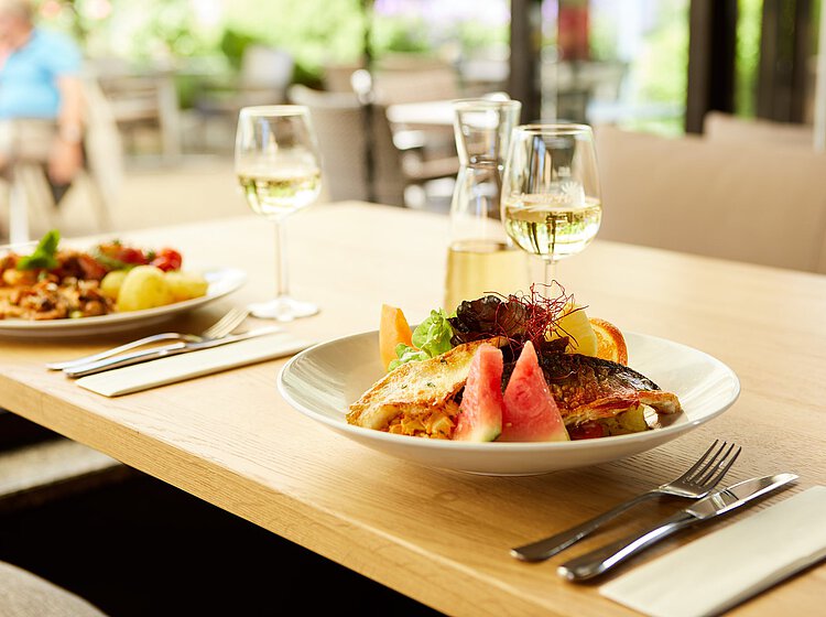 Ein gedeckter Tisch in einem Restaurant mit zwei Tellern, jeweils mit einer abwechslungsreichen Mahlzeit, dazu zwei Gläser Weißwein und eine Karaffe mit Getränk. Die Atmosphäre wirkt hell und einladend.