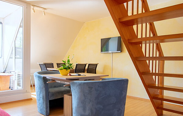 Heller Wohnbereich mit Holzwangentreppe, Esstisch mit gepolsterten Stühlen, Wand-TV und großen Fenstern, die Tageslicht hereinlassen.