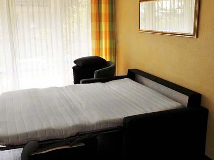 Schlafzimmer mit einem Bettsofa, das zu einem Bett mit weißer Matratze ausgezogen ist, neben einem Fenster mit lichtdurchlässigen Vorhängen.