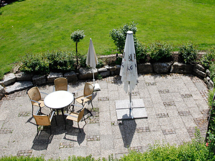 Terrasse Blick auf eine gepflasterte Terrasse mit einem runden Tisch, fünf Stühlen und zwei geschlossenen Sonnenschirmen, umgeben von grünem Rasen und Pflanzen.