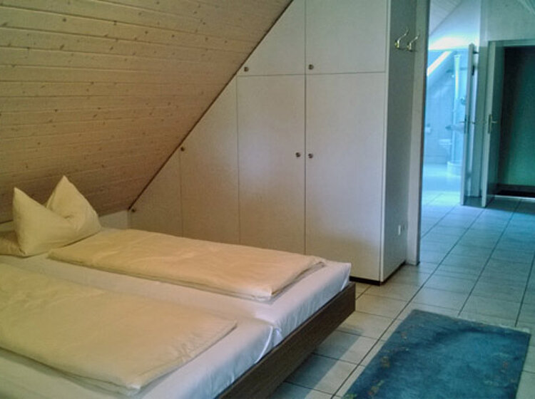 Schlafzimmer mit zwei Einzelbetten, schräger Holzwand, eingebautem Kleiderschrank und Blick in einen angrenzenden hellen Flur. Einfach möbliert, mit Kachelboden und Teppich.