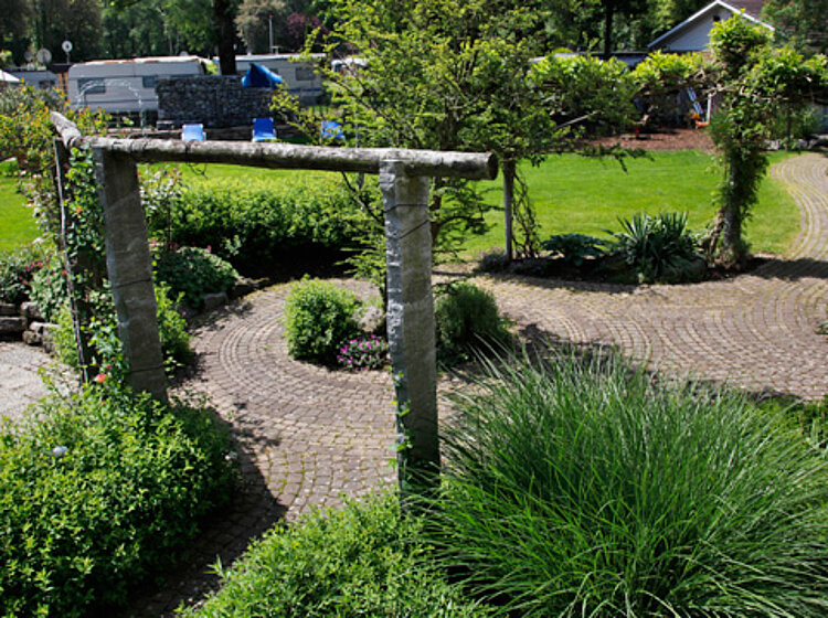 Gepflasterter Gartenweg Gepflasterter Gartenweg führt durch grüne Sträucher und Pflanzen, mit einem einfachen Holzbogen als Eingangselement. Sonniger Tag mit blühender Vegetation, ruhige und gepflegte Gartenanlage.