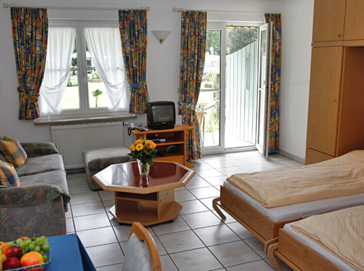 Helles Zimmer mit zwei Einzelbetten, Sofa, Couchtisch mit Blumen, Fernsehgerät, Fenster und geöffneter Terrassentür. Gemütlich und funktional eingerichtet.
