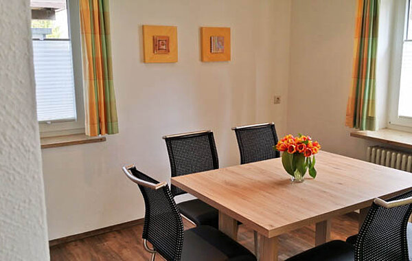 Moderner, heller Essbereich mit quadratischem Holztisch, vier schwarzen Stühlen und einer Vase mit oranger Blumen auf dem Tisch. Zwei Fenster mit Gardinen lassen Tageslicht herein. Weiße Wände sind mit zwei kleinen Bildern dekoriert.