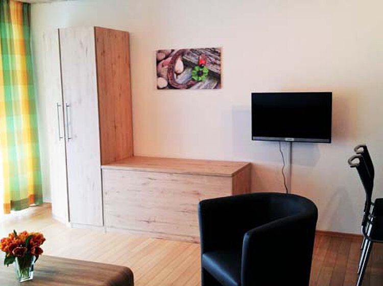 Helles, modern eingerichtetes Zimmer mit zwei schwarzen Sesseln, einem Tisch, einer Holzkommode, einem an der Wand befestigten Fernseher und einem Fenster mit gelb-grün karierten Vorhängen.